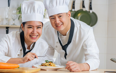 Two Asian professional couple chef wearing white uniform and hat, helping for preparing ingredients for healthy meal, cooking in kitchen together, smiling with happiness and confidence. Food Concept.