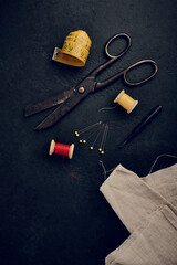 Sewing supplies flat lay - old tailors scissors, wooden thread spools, seam ripper and linen cloth, black background, space for text