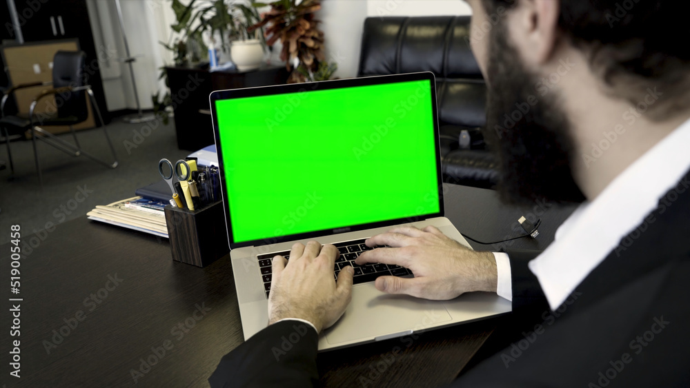 Poster close up for a bearded man typing on a computer laptop with a green screen. man in suit working in o