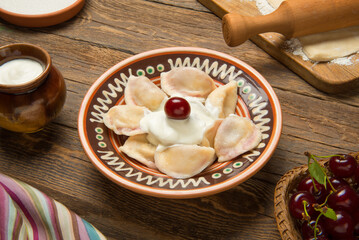 Traditional Ukrainian dumplings, vareniki with cherries in a ceramic painted bowl with ingredients on an old rural wooden table. - 519005882