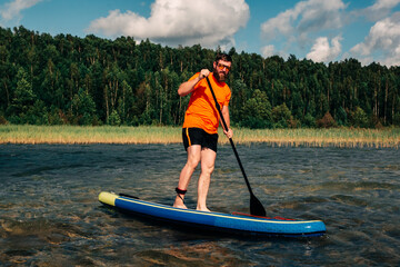 Fototapeta na wymiar Joyful red bearded man in sunglasses is training on a SUP board on a large lake during sunny day. Stand up paddle boarding - awesome active recreation in nature. 