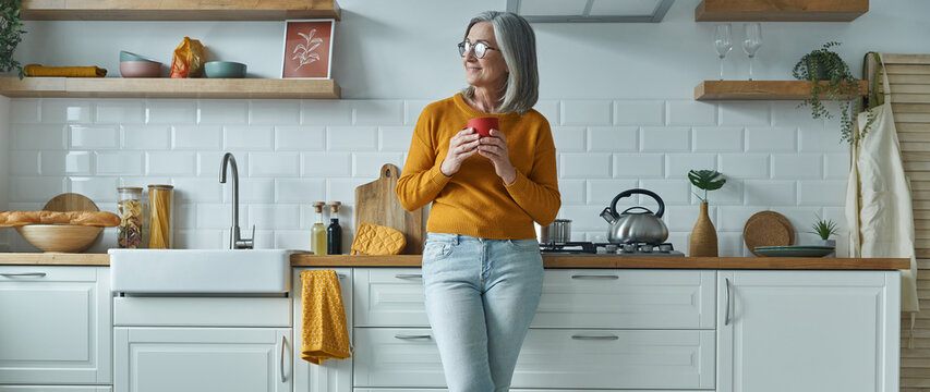 Thoughtful senior woman holding cup and smiling while standing at the domestic kitchen