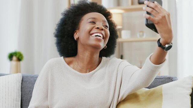 Black woman posing and taking selfies and making a peace hand gesture with her phone at home. One happy and cheerful female having fun taking photos for her social media while relaxing on the couch