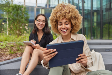 Two female groupmates rest outdoors after university lessons work on project together watch training webinar on tablet take notes in spiral notepad plan common coursework discuss creative solutions