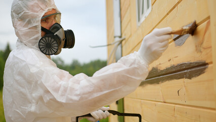 Man in respirator and special overall paints wooden house holding bucket with paint
