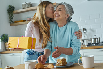 Young woman kissing her mother and holding a gift box while spending time at the kitchen together