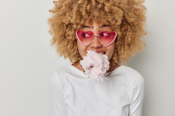 Horizontal shot of curly haired young woman has mouth full of cotton candy looks aside wears pink...