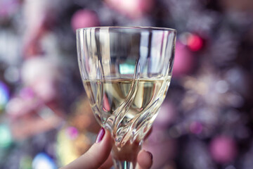 White wine glass in a female hand, woman holding a drink in front of a christmas tree, pink baubles, closeup, cheers, toast