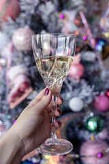 Christmas toast, white wine glass in a female hand, woman holding a drink in front of a tree, pink baubles, vertical, cheers