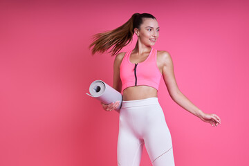 Beautiful young woman in sports clothing carrying an exercise mat against pink background
