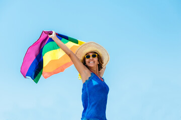 a woman in a straw hat, blue dress and yellow sunglasses, holding a rainbow flag on a blue sky background. lgtb concept and tolerance. Diversity