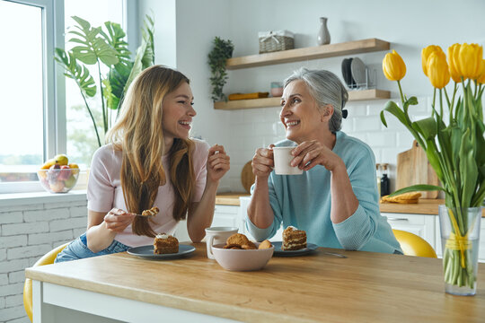 Senior mother and her adult daughter enjoying hot drinks and sweet food at the kitchen
