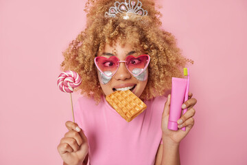 Indoor shot of funny curly haired woman holds lollipop waffle in mouth tube of toothpaste and toothbrush wears heart shaped sunglasses and t shirt isolated over pink background. Sugar addiction