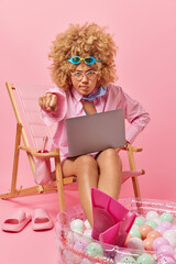 Obraz na płótnie Canvas Angry curly woman clenches fist looks irritated at camera wears goggles on forehead formal shirt and flippers works on laptop computer rests at beach poses on deck chair against pink background