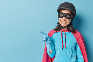Horizontal shot of happy woman dressed like superhero points away on blank space shows place for your advertisement isolated over blue background. Place your promotion here. Superpower concept