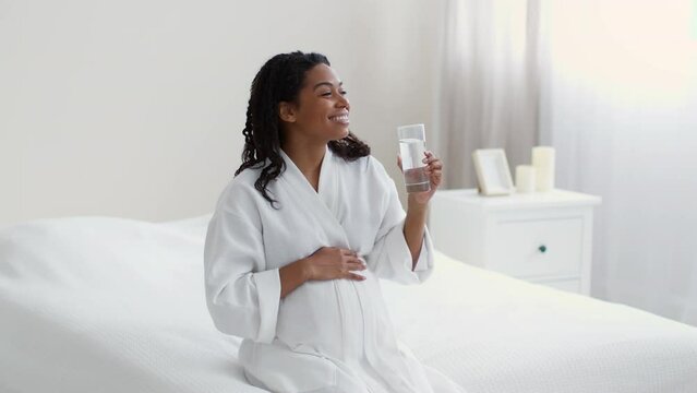 Water balance during pregnancy. Young expectant african american woman drinking water from glass and caressing belly