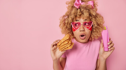 Horizontal shot of surprised curly haired woman wears casual t shirt feels stunned holds delicious handmade waffles toothpaste and toothbrush applies beauty patches under eye to moisturise skin