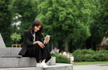 Charming woman in a business suit smiles and communicates with someone looking at the phone, sitting in the park on the stairs