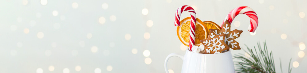 Hot winter drink in a white mug: cozy home composition with homemade gingerbread cookies, candy cane, fir tree branch. Wooden background, christmas lights and candles. Banner copy space