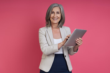 Happy senior woman in formalwear holding digital tablet while standing against pink background