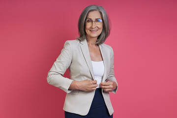 Confident senior woman buttoning her jacket while standing against pink background
