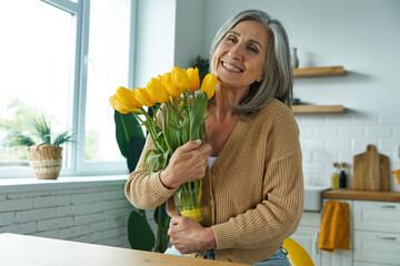 Happy senior woman holding a bunch of yellow tulips while standing at the domestic kitchen