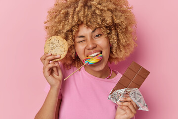 Photo of curly haired woman holds cookie bar of chocolate multicolored lollipop in mouth winks eyes dressed in t shirt enjoys eating sweet food isolated over pink background. Sugar addiction
