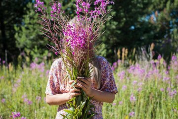 Young woman holding a bundle of pink fireweed flowers in front of her face. Girl hiding behind pink-violet wild flowers in summer nature.