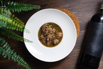 A portion of the Georgian dish chakapuli (young meat stewed with herbs and spices) in a plate seen from above. A plate on the table with a bottle of red wine next to it.