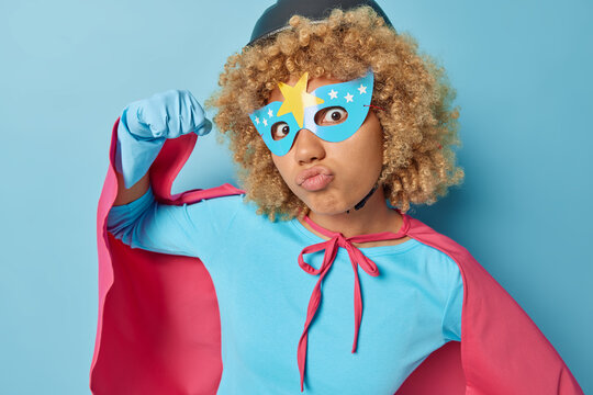 Powerful young woman with curly hair raises arm shows muscles pouts lips focused at camera prepares for masquerade dressed like superhero isolated over blue background. Feminism and superpower