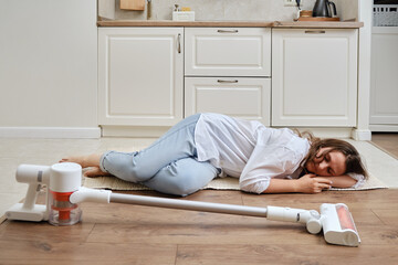 A tired woman with a wireless portable vacuum cleaner in the kitchen is lying on the floor. A...