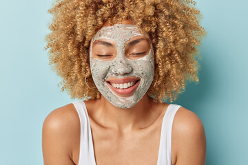 Portrait of cheerful woman smiles positively keeps eyes closed applies clay mask on face takes care of skin dressed in t shirt isolated over blue background. Facial procedures and everyday treatment
