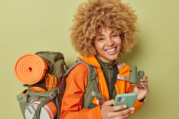 Horizontal shot of glad active woman camper uses mobile phone for chatting online holds binoculars carries heavy rucksack wears orange jacket isolated over dark green background. Tourism concept
