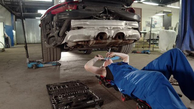 woman mechanic in car service rolls under sports car with wrench or ratchet in hands and repairs car. auto mechanic in blue overalls climbs under car and checks all details and reliability of engine.