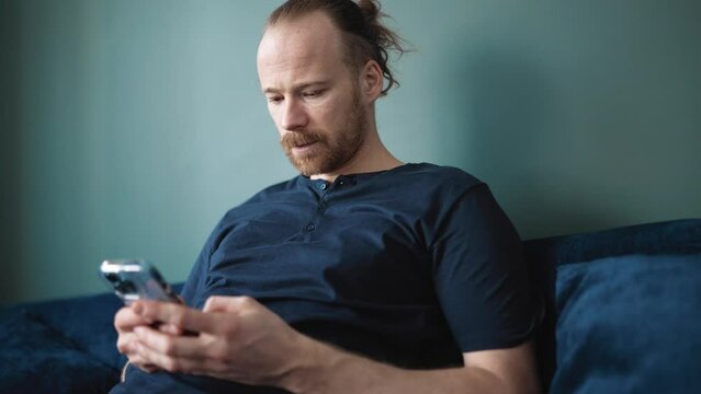 Laughing blond man wearing pajama texting on mobile while sitting on the sofa at home