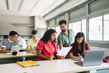Friendly Professor explaining exercise to students in high school classroom. Young Teacher with beard helping teenager girl