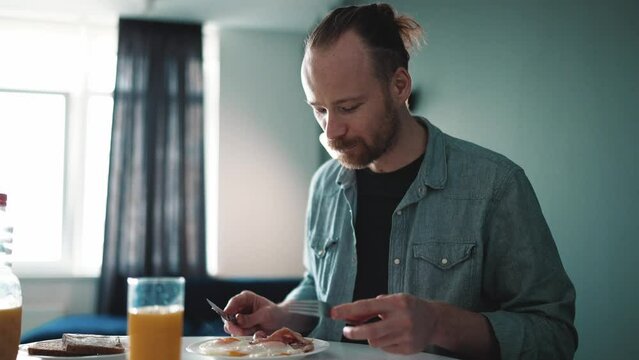 Positive blond man eating frying eggs with bacon in the morning at home