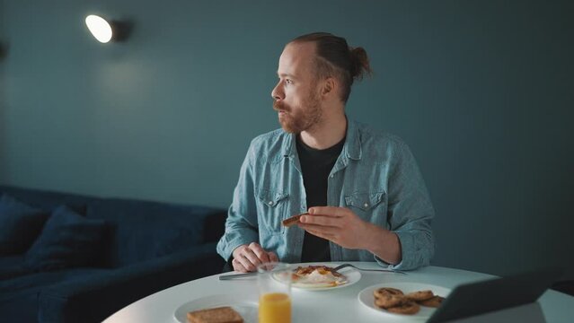 Concentrated blond man eating bread and looking around in the morning at home