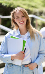 Smiling pretty happy blond girl university or college student holding notebooks looking at camera standing outside campus. Vertical portrait outdoors, education program courses and classes ads.