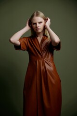 Expressive elegant young confident blonde lady in trendy stylish leather brown trench coat posing isolated touching hair holding hands overhead at studio green wall background. Fashion banner offer