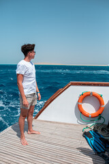 A handsome young guy in a white T-shirt and plaid shorts stands at the stern of the yacht and looks...