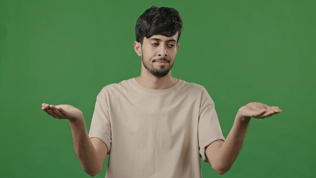 Puzzled man confused uncertain hispanic arab thoughtful young guy standing in green studio shrugs shoulders make choice decision choosing doing weights gesture with hands compares between two options