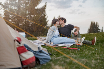 Happy man and woman kissing on grass at tent while resting at rural nature. Camping at wood.