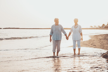 Couple of old mature people walking on the sand together and having fun on the sand of the beach enjoying and living the moment. Two cute seniors in love having fun. Barefoot walking on the water.