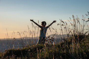 Mature woman in Punta Ballena, standing among weeds with her arms outstretched in front of the sea at sunset, with the Piriápolis hills in the background, in the department of Maldonado, Uruguay.
