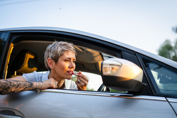 Naklejka premium One woman mature caucasian female businesswoman sitting in car putting lipstick fixing repairing makeup on her face while waiting in summer day evening real people copy space gray hair
