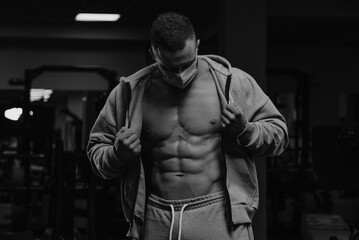 Fototapeta na wymiar A bodybuilder in a face mask to avoid the spread of coronavirus is opening his zipped hoodie to demonstrate his athletic physique. A sporty guy in a surgical mask is posing after a workout in a gym.
