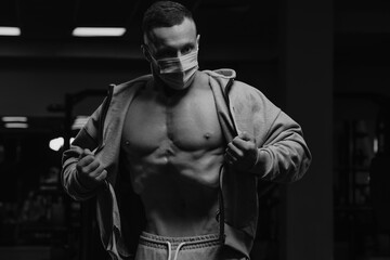 Fototapeta na wymiar A bodybuilder in a face mask to avoid the spread of coronavirus is opening his zipped hoodie to demonstrate his vacuum workout. A sporty guy in a surgical mask is posing after training in a gym.