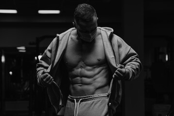 Fototapeta na wymiar A bodybuilder in a face mask to avoid the spread of coronavirus is opening his zipped hoodie to demonstrate his abdominal muscles. A sporty guy in a surgical mask is posing after a workout in a gym.