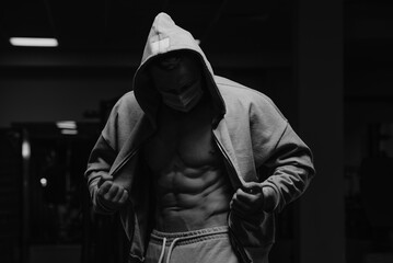 Fototapeta na wymiar A bodybuilder in a hood and a face mask to avoid the spread of coronavirus is opening his zipped hoodie to demonstrate his abdominal muscles. A sporty guy in a surgical mask is posing after a workout